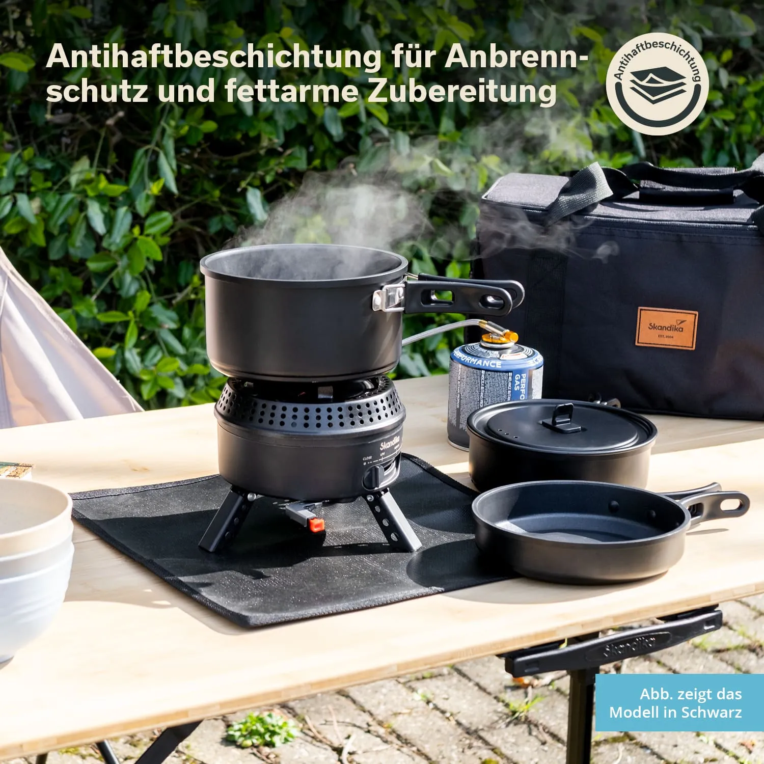 Kochgerät mit Antihaftbeschichtung auf einem Tisch im Freien. Dampf steigt aus einem Topf auf. Weitere Kochtöpfe sind sichtbar.