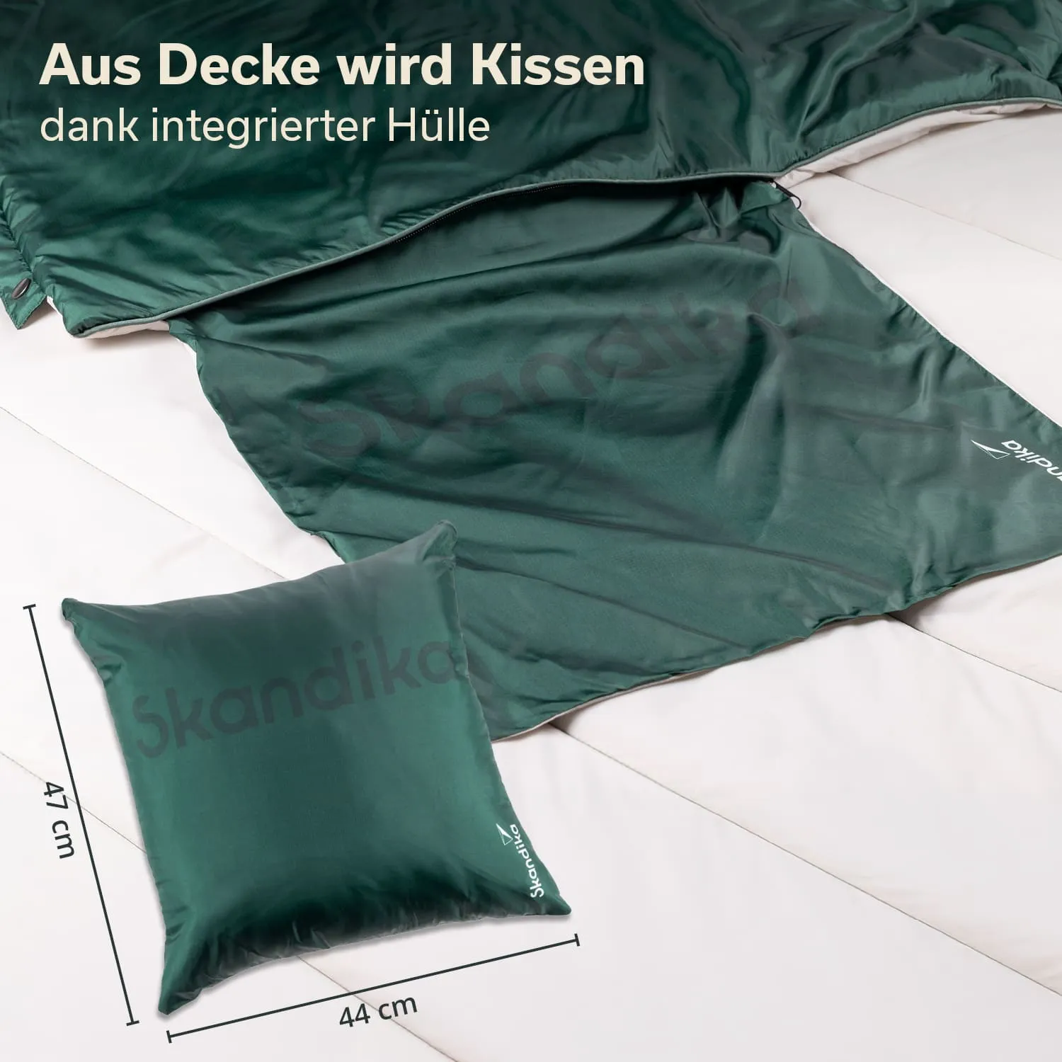Grüne Decke und Kissen auf einer hellen Oberfläche. Kissen misst 44 cm x 47 cm. Auf der Decke ist der Schriftzug „Skandika“.