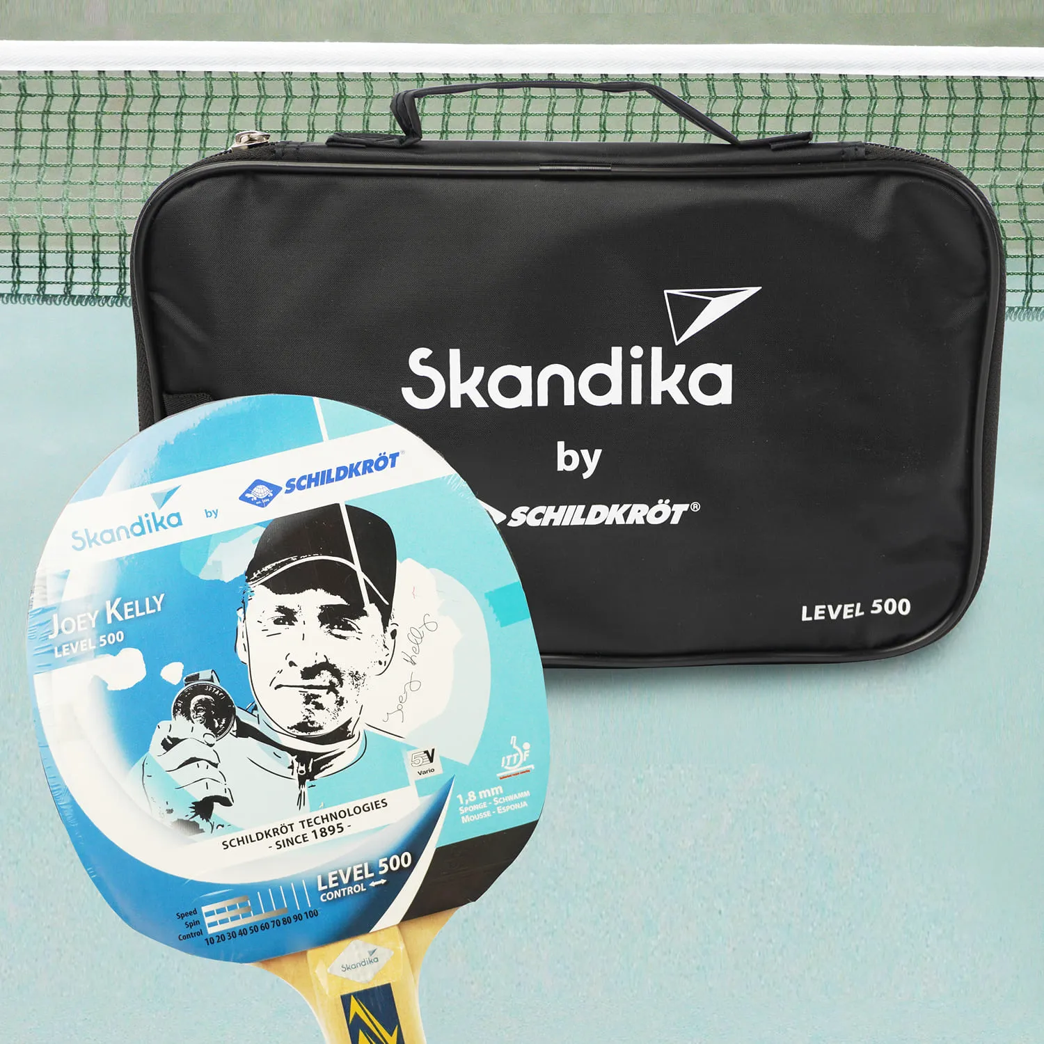 Tischtennisschläger mit einer blauen Oberfläche und der Aufschrift „Joey Kelly LEVEL 500“ neben einer schwarzen Tasche mit dem Logo „Skandika by Schildkröt“. Im Hintergrund ist ein Tischtennisnetz zu sehen.