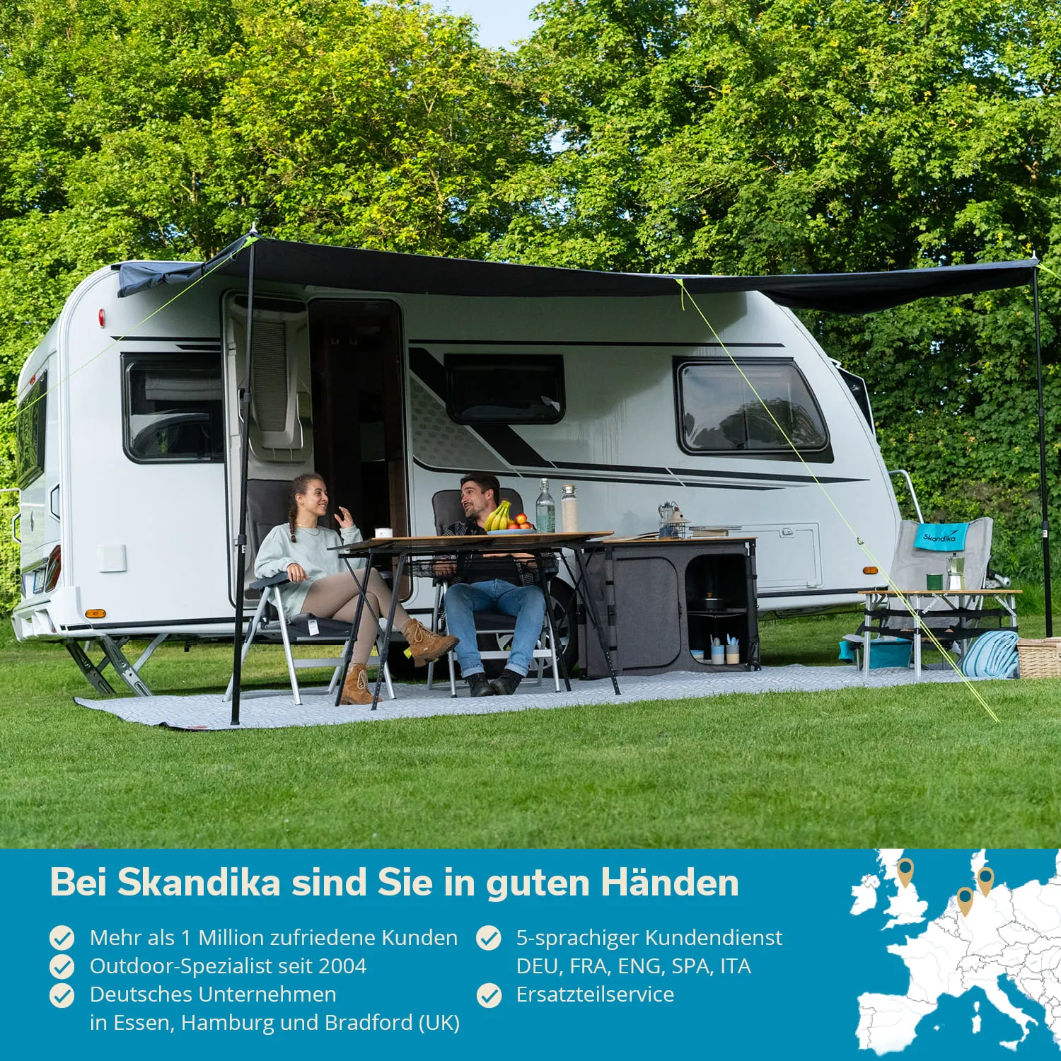 Wohnmobil auf einer Wiese mit zwei Personen am Tisch. Umgeben von Bäumen. Tisch mit Obst und Getränken.