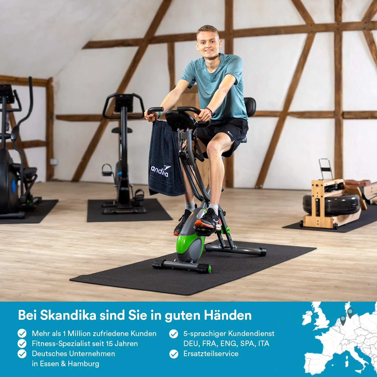 Eine Person sitzt auf einem Heimtrainer in einem Raum mit Holzdecken. Im Hintergrund sind weitere Fitnessgeräte sichtbar.