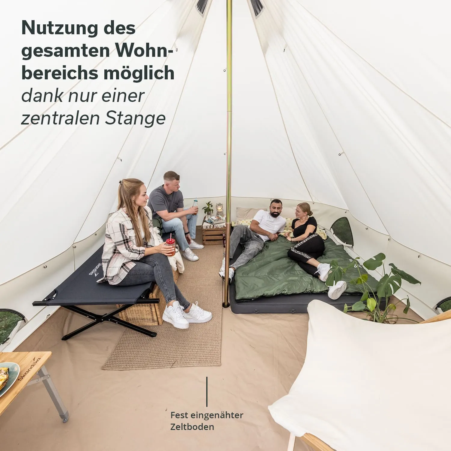 Zeltinnenraum mit einer zentralen Stange. Vier Personen sitzen und stehen auf verschiedenen Schlafgelegenheiten. Möbel und Pflanzen sind sichtbar.