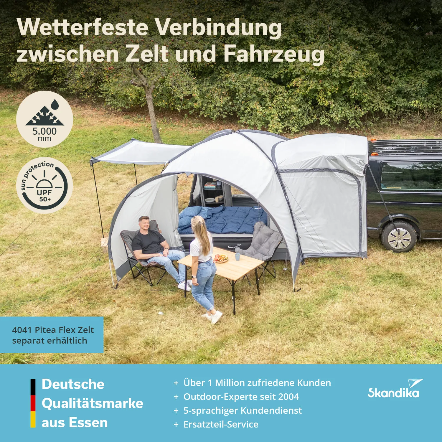Zelt mit Anbau, Verbindung zu einem Fahrzeug. Zwei Personen sitzen an einem Tisch. Grüne Wiese, Bäume im Hintergrund.
