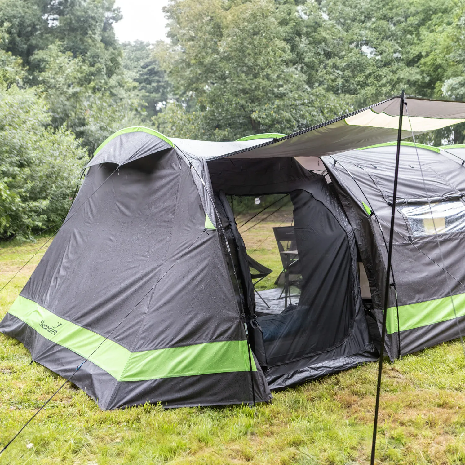 Zelt in dunklem Grau mit grünen Streifen, in einer Wiese zwischen Bäumen. Vor dem Zelt ist ein kleiner Tisch mit Stühlen.