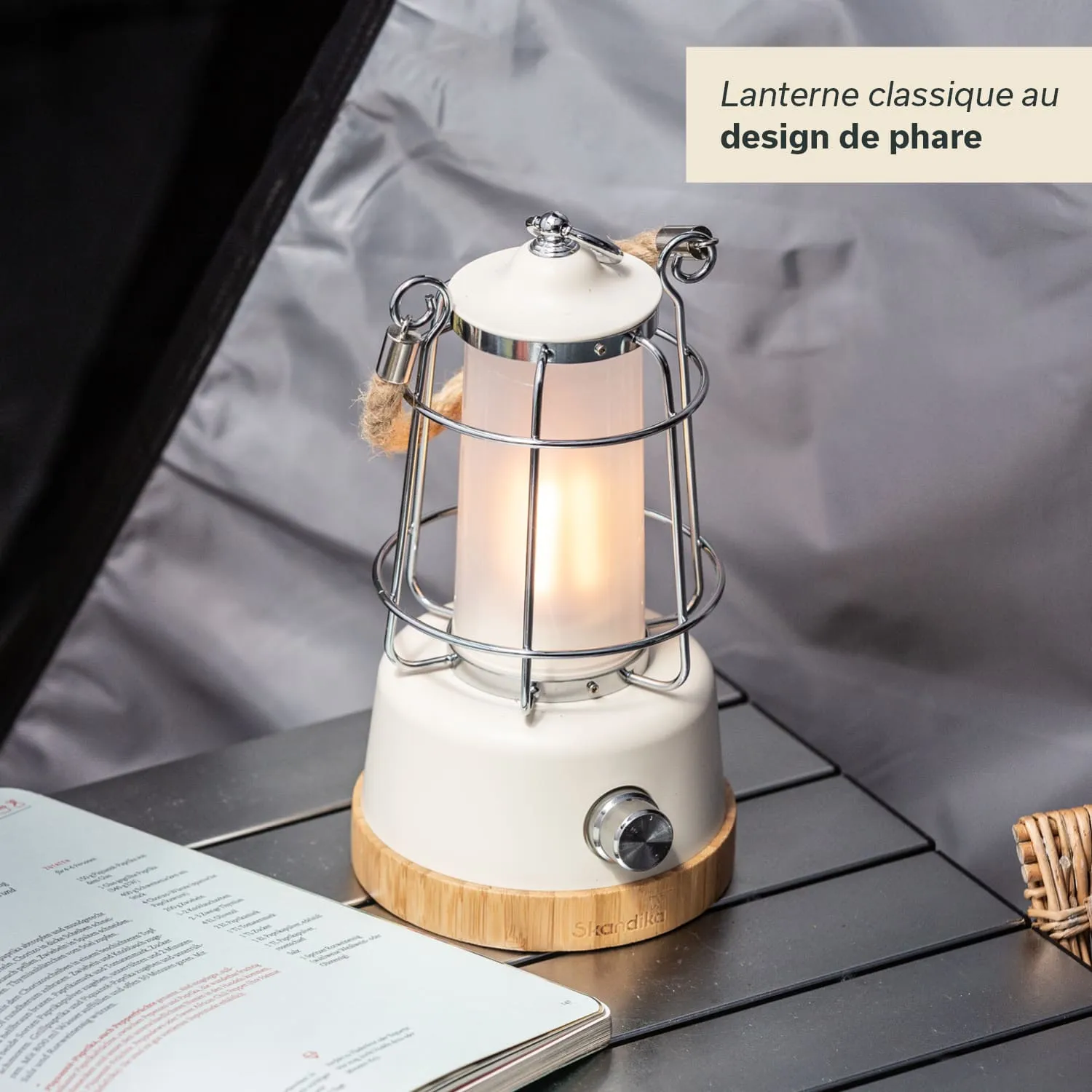 Eine klassische Laterne im Design eines Leuchtturms steht auf einem Tisch. Die Laterne hat einen weißen, zylindrischen Körper mit einem Metallgitter und einem Holzsockel. Daneben liegt ein aufgeschlagenes Buch.