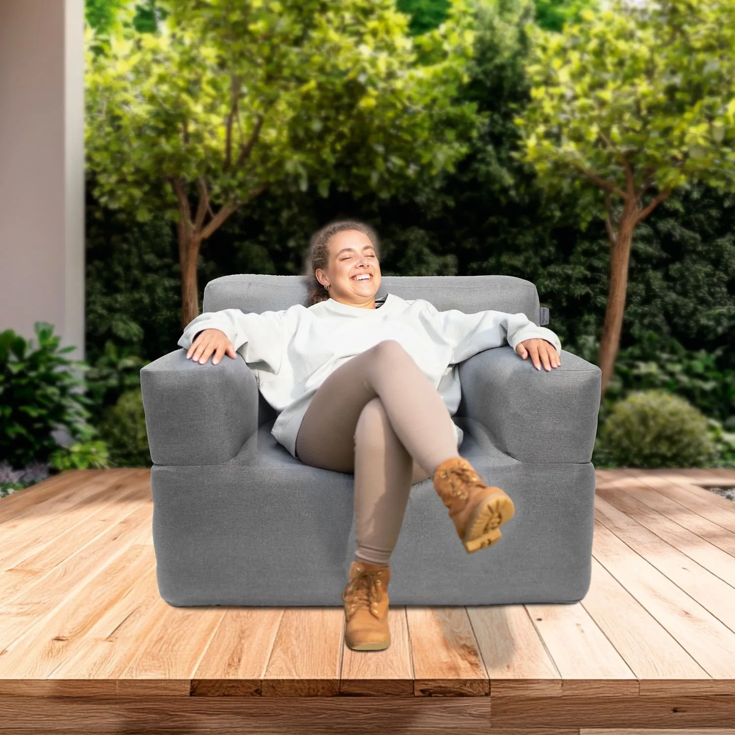 Eine Person sitzt lächelnd auf einem grauen Sofa. Der Hintergrund zeigt grüne Bäume und Pflanzen. Der Boden ist aus Holz.