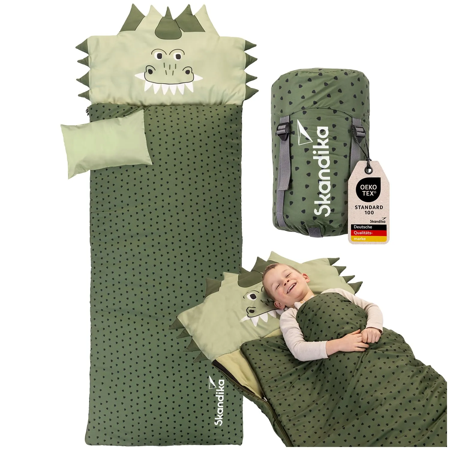 Schlafsack in Grün mit Drachenkopf und Punkten, daneben ein gerollter Schlafsack und ein Schild mit Pflegehinweisen. Ein Kind liegt lächelnd darin.