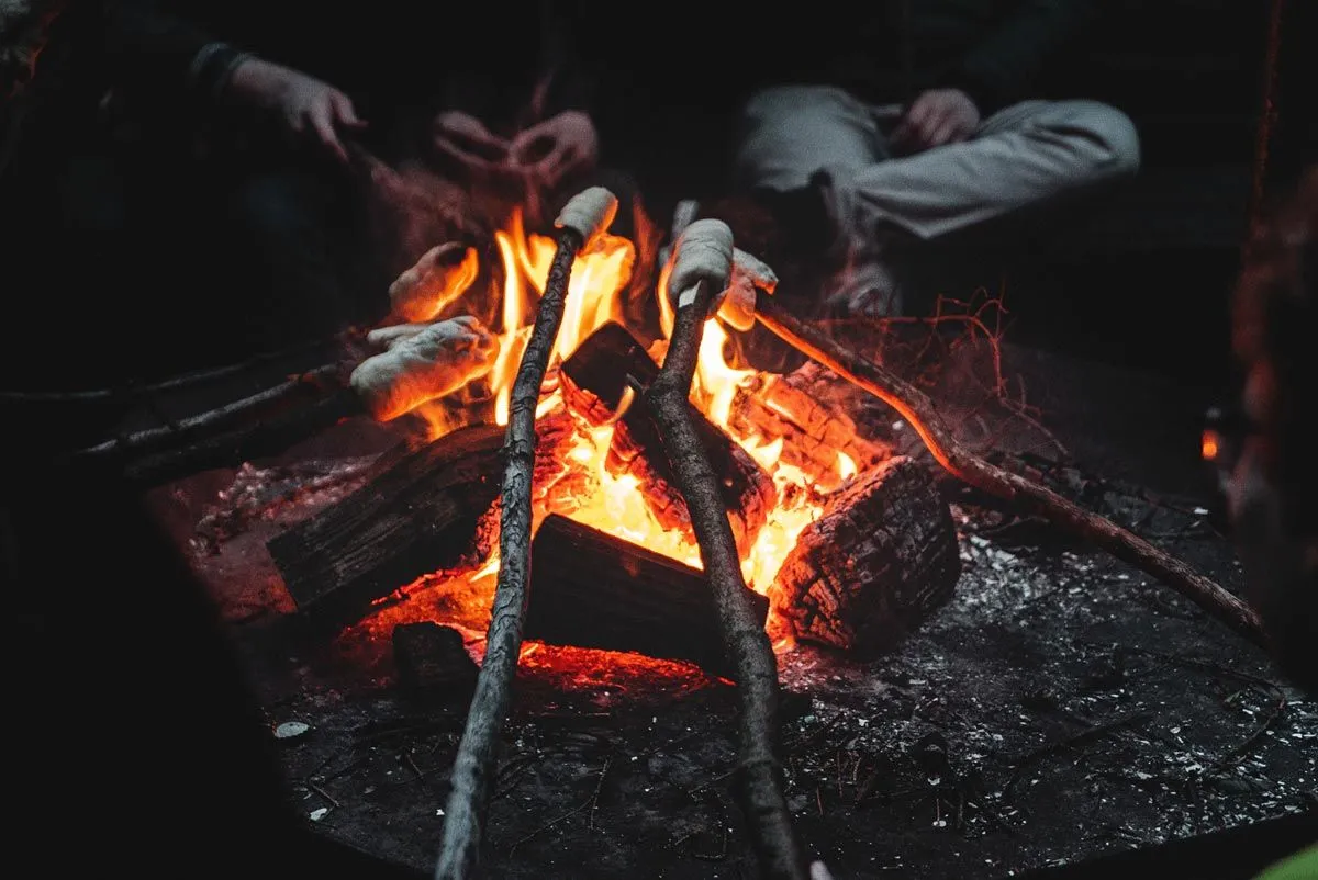 Stockbrot über einem Lagefeuer im Dunkeln mit Person um das Feuer sitzend