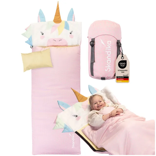 Schlafsack mit Einhorn-Motiv und pinkem gepunktetem Design. Eine Person liegt lächelnd darin. Ein pinker Rucksack ist daneben sichtbar.