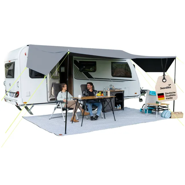 Ein Wohnwagen mit grauem Vordach, davor stehen ein Tisch und zwei Stühle. Auf dem Tisch sind Getränke und Obst. Zwei Personen sitzen entspannt.