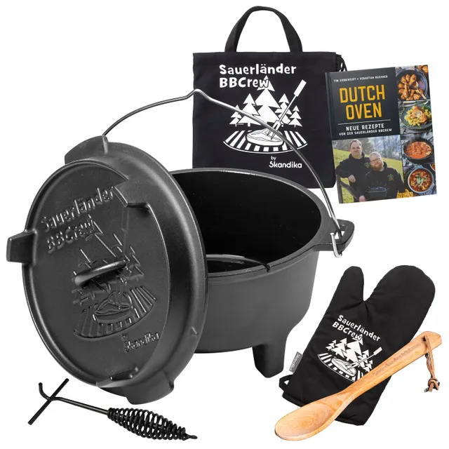 Ein Dutch Oven aus Gusseisen mit Deckel, ein schwarzes Trageband, ein Kochbuch, zwei Ofenhandschuhe und ein Holzlöffel.