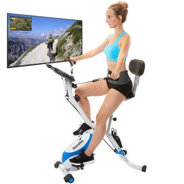 Eine Person sitzt auf einem Heimtrainer mit Armlehnen. Im Hintergrund ist ein großer Bildschirm mit einer Fahrradstrecke in der Natur zu sehen.