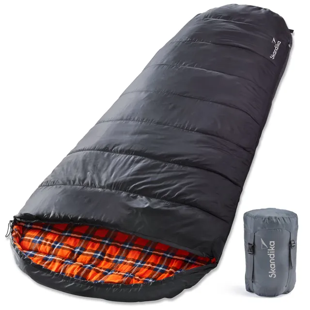 Schlafsack in Schwarz mit kariertem Innenfutter in Orange und einem grauen Packbeutel daneben.