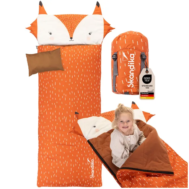 Ein orangefarbener Schlafsack mit Fuchs Kopfteil und Muster. Daneben ein Rucksack in ähnlicher Farbe. Eine Person sitzt im Schlafsack.