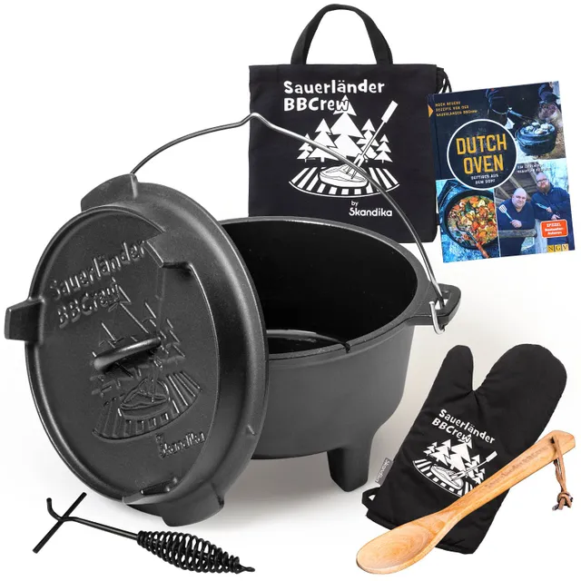 Dutch Oven aus Gusseisen mit Deckel, neben einem Kochbuch, einer Tragetasche, einem Holzlöffel und einem Topflappen.